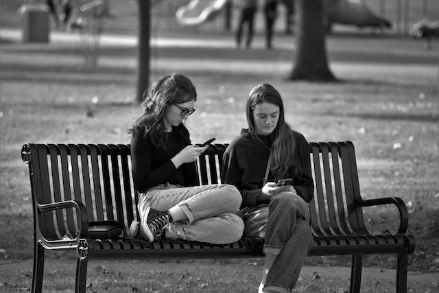 Two young women staring at their smartphones on a park bench