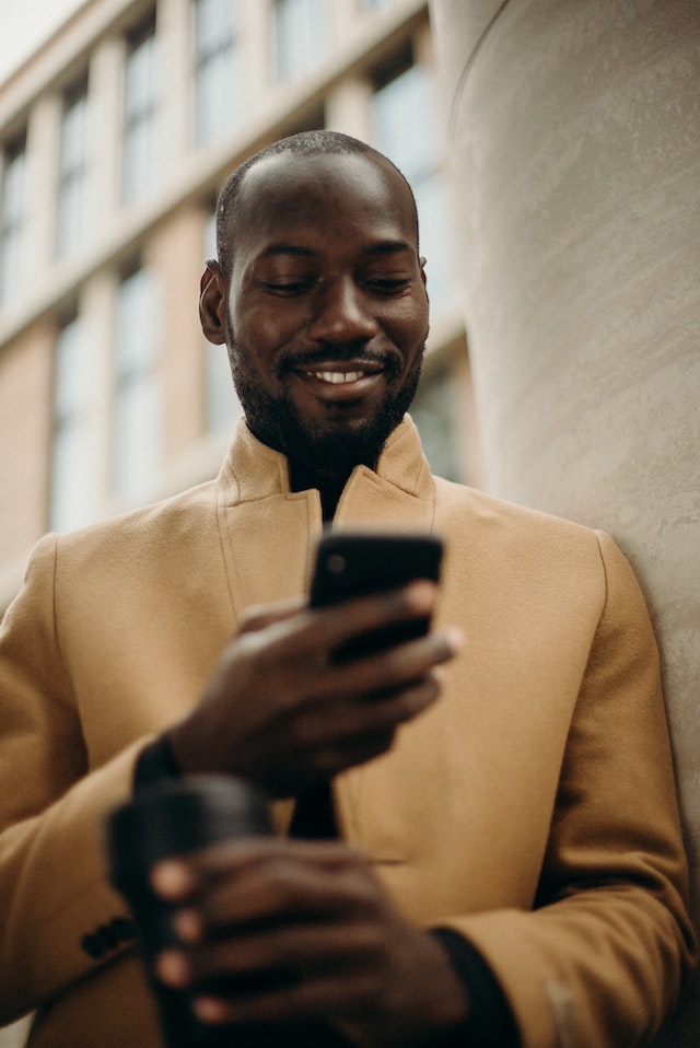 Man in tan coat smiling down at phone in his hand
